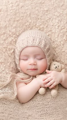 a baby sleeping with a teddy bear on top of it's arm and wearing a knitted hat