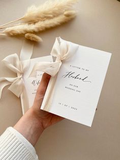 a person holding a wedding card in their left hand with a ribbon around it and some feathers on the table