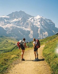 two people with backpacks walking up a dirt path in front of snow capped mountains