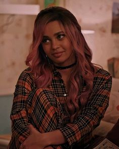 a woman with pink hair is sitting on a couch and looking at the camera while holding her arms crossed