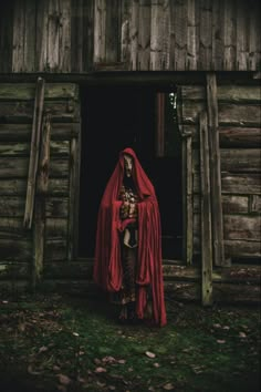 a woman in a red cape and dress standing outside of a wooden building with a cat on her chest