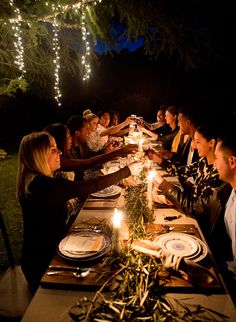 a group of people sitting at a long table with plates and candles on it in the dark