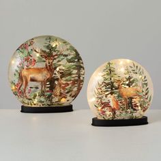 two snow globes with deer and pine trees on them, one is illuminated by fairy lights