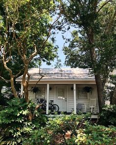 a small white house surrounded by trees and bushes with two chairs on the front porch