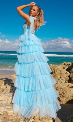 a woman in a blue dress standing on rocks near the ocean with her arms behind her head