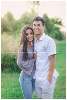 a young man and woman posing for a photo in front of some grass with trees