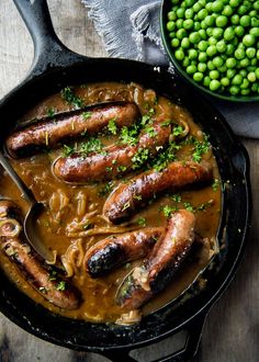 some sausages and noodles in a skillet next to peas on a wooden table