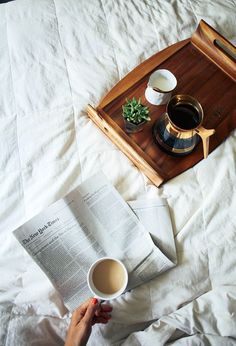 a person is reading a book and holding a cup of coffee on a bed with white sheets