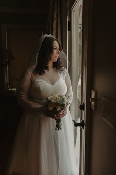 a woman standing in front of a door wearing a wedding dress and holding a bouquet