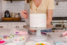 a woman standing in front of a cake on top of a white counter next to bowls and utensils