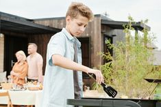 a young boy grilling food on an outdoor grill with family in the back ground