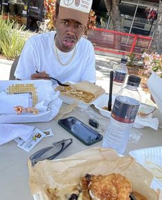 a man sitting at a table with food and drinks in front of him, wearing a hat