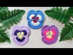 three crocheted flowers sitting next to each other on top of a table with green leaves