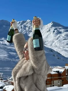 a woman holding two bottles of champagne in the snow with mountains in the back ground