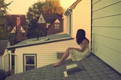 a woman sitting on top of a roof next to a house and looking at the sky