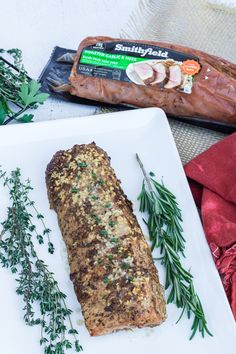 a piece of meat sitting on top of a white plate next to a knife and some herbs