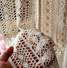 a hand holding a piece of crocheted fabric in front of a window curtain