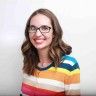 a woman wearing glasses and a colorful sweater smiles at the camera while standing in front of a white wall