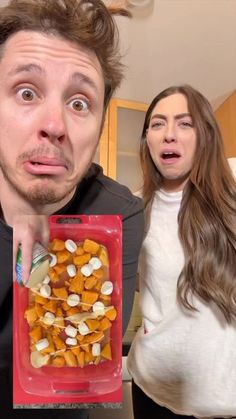 a man and woman standing in front of a plastic container filled with food, both looking surprised at the camera