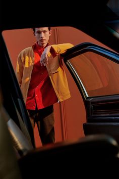 a man standing next to a car with his hand on the door handle and wearing a red shirt