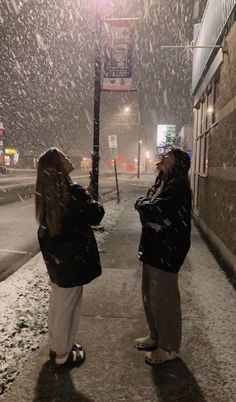 two women standing in the snow on a street corner looking up into the sky at something