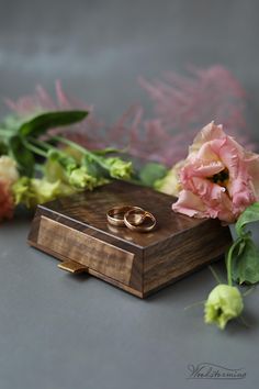 two gold wedding rings sitting on top of a wooden box next to pink and green flowers