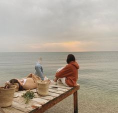 two people sitting on a wooden table by the water with baskets and food in front of them