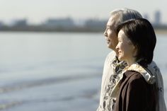 an older couple standing next to each other on the beach looking out at the water