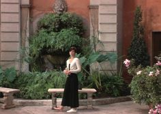 a woman standing in front of a building with flowers and plants around her, next to a bench