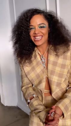 a woman with long curly hair wearing a tan suit and silver necklace smiling at the camera