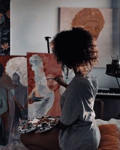 a woman sitting on the floor in front of some paintings and holding a paintbrush