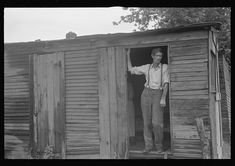 an old photo of a man standing in the open door of a small wooden cabin