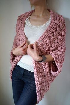 a woman wearing a pink crocheted shawl and white top standing in front of a wall