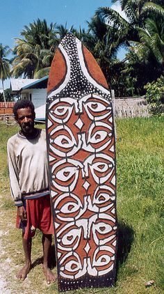 a man standing next to a surfboard on top of a grass covered field with palm trees in the background