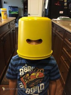 a young boy wearing a large yellow plastic bucket on his head in a kitchen area