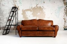 a brown leather couch sitting in front of a wall with peeling paint and a ladder leaning against it