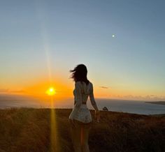 a woman standing on top of a grass covered hillside next to the ocean at sunset