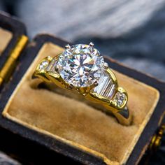 a diamond ring sitting on top of a velvet box with gold trimmings and two side stones