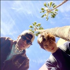 two young men standing next to each other under a palm tree on a sunny day