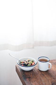 a bowl of food next to a cup of coffee on a wooden table in front of a window