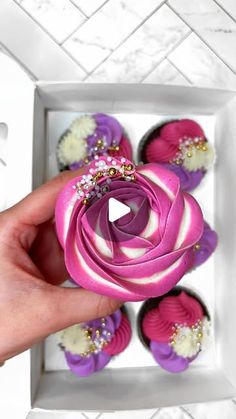 a person is holding a pink and purple flower in a box with cupcakes