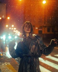 a woman standing in the rain with her hands up to her chest and wearing a black trench coat