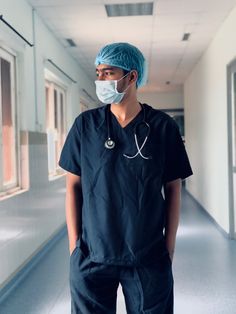 a man wearing scrubs and a surgical mask stands in a hospital hallway with his hands on his hips