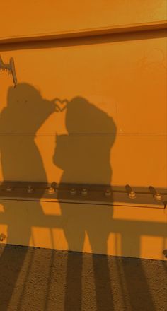 the shadow of two people on a yellow wall next to a metal bench and rail