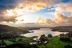 an aerial view of a lake surrounded by green fields and mountains under a cloudy sky