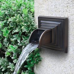 a water fountain spewing out from the side of a stone wall next to green bushes