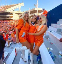 three women dressed in orange posing for a photo at a football game with the crowd behind them