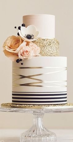 a three tiered cake decorated with flowers and gold glitters on a glass stand