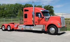 a red semi truck parked in a parking lot next to a chain link fence and trees