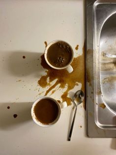 two cups of coffee sit in front of a dirty metal tray with spoons on it